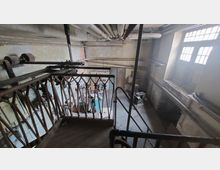 Ein verlassener, industriell anmutender Raum mit einer Metallbalustrade im Vordergrund, freiliegenden Rohren an der Decke und großen Fenstern, die diffuses Licht einlassen. Im Hintergrund sind alte Maschinen und Werkzeuge erkennbar, die auf einen früher genutzten Arbeitsraum hindeuten.