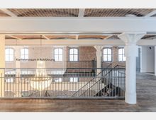 Industrie-inspiriertes Gebäudeinterieur mit Backsteinwänden, großen Sprossenfenstern und weißen Säulen. Im Vordergrund befindet sich ein Geländer, dahinter eine Treppe und ein großer, funkelnder Kronleuchter.