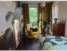 Gemütliches Schlafzimmer mit großer Wald-Fototapete an der linken Wand, dunklem Holzboden und einem gelben Ohrensessel neben einem kleinen Beistelltisch. Vor bodentiefen Fenstern mit goldenen Vorhängen stehen eine hohe Zimmerpflanze und eine Holzkommode; im Vordergrund ist ein Bett mit gemusterter Decke zu sehen.