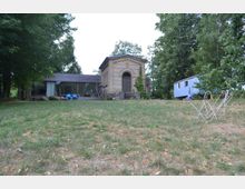 Eine weitläufige Wiese mit einem Gebäude aus gelbem Klinkerstein im Zentrum, das an eine historische Kapelle erinnert, daneben ein moderner Anbau mit Glasfassade. Im Hintergrund stehen Bäume, rechts ist ein blauer Bauwagen zu sehen, und im Vordergrund stehen mehrere helle Wäschespinnen.