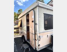 Ein älterer Wohnwagen steht auf einem Parkplatz mit geöffneter Eingangstür. Der cremefarbene Aufbau mit abgerundeten Kanten und Fenstern auf der Seite zeigt leichte Gebrauchsspuren, daneben ist eine kleine Tritthilfe aufgestellt.