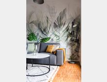 Wohnzimmer mit moderner Einrichtung: Ein graues Sofa mit bunten Kissen und einer orangenen Decke steht vor einer Wand mit großflächigem Blattmotiv. Im Vordergrund ein runder Couchtisch und ein Teppich auf hellem Holzfußboden, rechts eine schwarze Stehlampe.