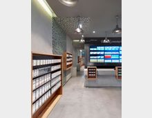 Innenansicht einer modern gestalteten Apotheke mit grauem Boden, hölzernen Regalen, die mit Produkten gefüllt sind, und einer digitalen Wand mit farbigen Anzeigen. Industrial-Design-Lampen hängen von der Decke, die teils mit einem Mosaikmuster verkleidet ist.