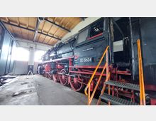 Historische Dampflokomotive in einer Werkstatthalle mit Holzbalkendecke, großen Fenstern und Schiebetoren. Im Vordergrund führt eine metallene Treppe mit orangefarbenem Geländer zur Lokomotive.