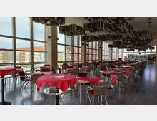 Großer, lichtdurchfluteter Speisesaal mit deckenhohen Fenstern und rot gedeckten Tischen. Der Raum ist mit Holzstühlen ausgestattet, die Decke zeigt auffällige hängende Akustikelemente, und im Hintergrund befindet sich eine Theke.