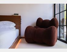 Ein modernes Schlafzimmer mit einem hölzernen Bettgestell, weißen Bettbezügen und einem braunen, gepolsterten Sessel mit organischen Formen. Rechts im Bild befindet sich ein großes Fenster mit schwarzem Metallrahmen, das Tageslicht hereinlässt.