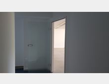 Ein heller Innenraum mit einer geöffneten Glastür, die in einen leeren Raum mit weiß gestrichenen Wänden und einer glatten, grauen Bodenfläche führt. Im Hintergrund sind an der Decke rechteckige Lampen und Kabelkanäle sichtbar.