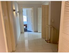 Flur in einem Wohnhaus mit glänzendem, hellen Fliesenboden, weißen Wänden und modernen Wandleuchten. Am Ende des Flurs ist eine offene Tür, die ins Schlafzimmer führt, rechts befindet sich ein Treppenabgang mit einem weißen Geländer.