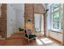 Wohnraum mit hellen Holzböden, freiliegenden Ziegelwänden und einem großen Fenster, das auf einen begrünten Außenbereich blickt. Im Vordergrund steht ein Sessel mit bunter Decke neben einer Stehlampe und einer Pflanze, während im Hintergrund ein Esstisch mit grünem Stuhl sichtbar ist.