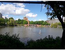 Ein ruhiger See in einer städtischen Parklandschaft, umgeben von Bäumen und Gebäuden. Im Hintergrund ist eine Backsteinkirche mit grüner Kuppel zu sehen, und kleine Wasserfontänen befinden sich im See.