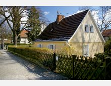 Gelbes Einfamilienhaus mit Satteldach und weiß gerahmten Fenstern, umgeben von einem Garten mit Holzzaun und Hecken. Im Vordergrund führt ein gepflasterter Gehweg entlang des Grundstücks, umrahmt von hohen Bäumen.