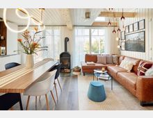 Modernes Wohnzimmer mit offenen Wohn- und Essbereich, einer großen cognacfarbenen Ledercouch, Holzbalkendecke, Kaminofen und einem Esstisch mit schwarzen und weißen Stühlen. Große Fenster mit weißen Vorhängen lassen viel Tageslicht herein, und Pendelleuchten sowie Bilder an der Wand ergänzen das gemütliche Ambiente.