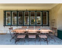 Überdachte Terrasse mit einem großen Holztisch und mehreren Klappstühlen aus Holz und Metall. Im Hintergrund befinden sich vergitterte Fenster und eine helle Putzwand, rechts lehnt eine Holzleiter an der Wand.
