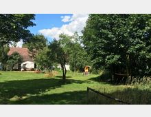 Grüne Wiese mit vereinzelten Bäumen, dahinter ein Haus mit rotem Ziegeldach und weißer Fassade. Am rechten Bildrand steht ein kleines, rundes Holzspielhäuschen, umgeben von Bäumen und Schatten.