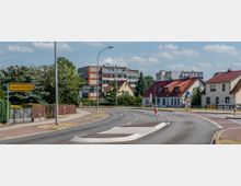 Stadtschlängelnder Straßenabschnitt mit Fußweg, umgeben von Einfamilienhäusern mit roten Dächern, einem mehrstöckigen Wohngebäude im Hintergrund und einem Wegweiser, der den Ort „Beeskow“ in 30 km Entfernung anzeigt.