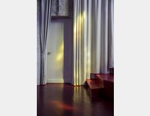 Ein Innenraum mit roten, glänzenden Böden, einer geschlossenen weißen Tür und langen, weißen Vorhängen, die teilweise einen Treppenaufgang verdecken. Farbige Lichtreflexionen in Gelb und Rosa sind auf dem Boden und den Vorhängen sichtbar.