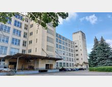 Großes, beige-farbenes Industriegebäude mit mehreren Etagen und zahlreichen Fenstern, links ein Ladebereich mit Überdachung. Im Vordergrund ein Parkplatz mit Autos, rechts davon hohe Nadelbäume.