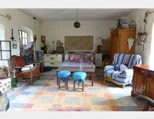 Ein gemütlich eingerichteter Wohnraum mit rustikalem Charme, ausgestattet mit einem Bett, zwei gepolsterten Hockern, einem gestreiften Sessel und einem antiken Holzschrank. Der Raum hat gemusterte Fliesen, weiße Wände, ein großes Fenster und eine Mischung aus dekorativen Vintage-Elementen.