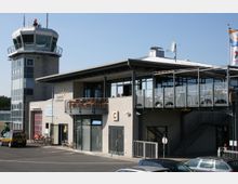 Flughafengebäude mit einem Kontrollturm links und einem modernen Hauptgebäude, das große Fenster, Balkone und eine Außenterrasse mit Sitzgelegenheiten zeigt. Im Vordergrund sind parkende Autos, Fahrräder und ein Flughafenfahrzeug sichtbar.