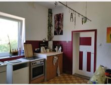 Eine einfache Küche mit weißen Wänden und rot-weißen Akzenten. Sichtbar sind ein Herd mit Backofen, eine Arbeitsplatte aus Holz, eine Spüle sowie Regale mit Flaschen und Küchenutensilien. Ein großes Fenster auf der linken Seite lässt Tageslicht herein.