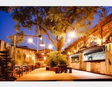 Beleuchteter Außenbereich einer Bar mit Holztischen und -stühlen auf einer Holzterrasse, umgeben von Pflanzen und einem großen Baum. Im Hintergrund befinden sich Backsteinfassaden mit Graffiti, daneben Verkaufsstände mit rustikaler Gestaltung.