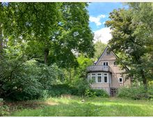 Ein zweistöckiges Holzhaus mit Satteldach und einer kleinen Terrasse, umgeben von dichtem Grün aus Bäumen, Sträuchern und einer Wiese. Der Himmel ist teils bewölkt, und das Grundstück wirkt wie in einem ländlichen oder waldreichen Gebiet gelegen.