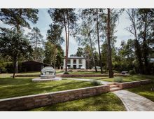 Modernes Haus mit weißen Fassaden und Balkonen, umgeben von hohen Bäumen in einer parkähnlichen Umgebung. Im Vordergrund gepflegter Rasen mit gepflasterten Wegen, Terrassen aus Naturstein und einer runden Gartenliege.