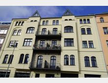 Fassade eines mehrstöckigen Wohngebäudes mit hellgelbem Putz und einer Mischung aus großen rechteckigen und bogenförmigen Fenstern. Zwei Etagen verfügen über Balkone mit Geländern und Pflanzentöpfen, das Dach ist mit gläsernen Spitzgauben gestaltet.