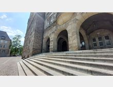 Steinerner Eingang eines historischen Gebäudes mit breiter Treppe und mehreren Rundbogenarkaden. Rechts sind Fenster mit Gitterstäben, links ein kleiner Platz mit Bäumen und umliegenden Gebäuden.