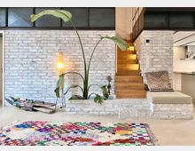 Wohnbereich mit weiß gestrichener Ziegelwand, Holztreppe mit integrierter Beleuchtung, großer Topfpflanze und moderner Stehleuchte. Im Vordergrund liegt ein bunter Teppich, während rechts ein Sitzbereich mit Kissen zu sehen ist.