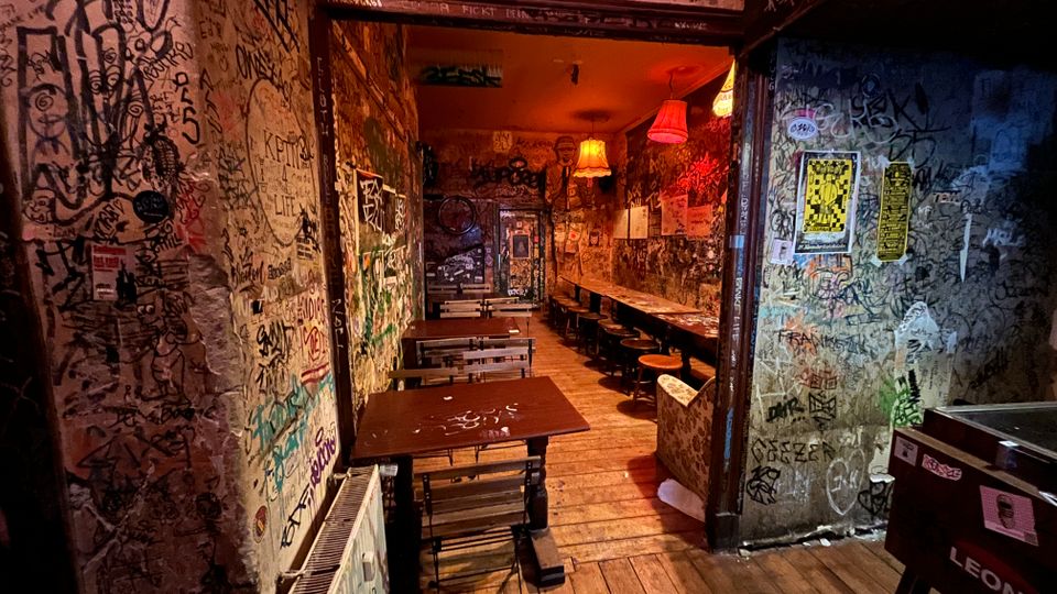Schmaler Innenraum einer Bar oder eines Cafés mit Holzboden, einfachen Tischen und Hockern entlang einer langen Theke. Wände, Türrahmen und Decke sind dicht mit Graffiti und Aufklebern bedeckt, beleuchtet von warmen, hängenden Lampenschirmen.