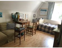 Ein gemütliches Wohn- und Schlafzimmer mit hellem Holzboden, einem karierten Sofa, einer Tagesliege mit gemusterten Decken und einem kleinen Holztisch mit Tischlampe und einem Globus. An der Wand hängen Bilder, und ein Fenster mit hellblauen Vorhängen lässt Licht herein.