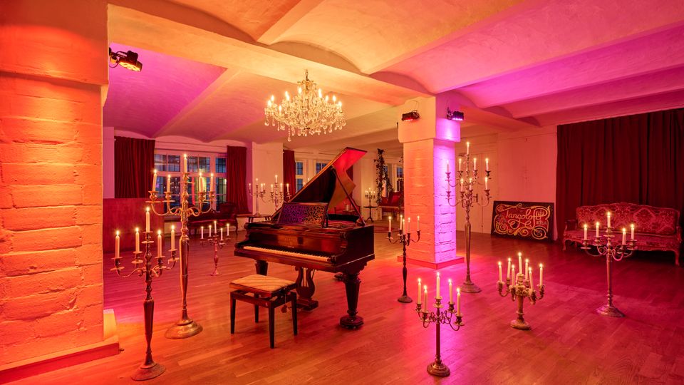 Eleganter Veranstaltungsraum mit Holzboden, weißen Stützsäulen und gewölbter Decke, in warmem Rot‑ und Violettlicht ausgeleuchtet. In der Mitte steht ein Flügel, umgeben von mehreren Kerzenleuchtern und einem Kronleuchter; im Hintergrund Fenster mit Vorhängen und eine Sitzecke.