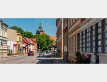 Straßenszene in einer Altstadt mit zweispuriger Fahrbahn, Gehweg und dicht stehenden, teils farbigen Gebäudefassaden mit Satteldächern. Im Hintergrund ragen Bäume und der Turm einer Kirche über die Häuser, entlang der Straße stehen Verkehrsschilder und einige geparkte bzw. fahrende Autos.
