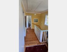 Schmaler Flur in einem Wohnhaus mit gelb gestrichenen Wänden, Holzdielenboden und weißer Decke mit einfacher Hängelampe. Rechts führt ein Treppenabgang mit weißem Geländer nach unten, daneben ein Fenster; am Ende des Flurs ist eine offene Tür zu einem angrenzenden Raum sichtbar.