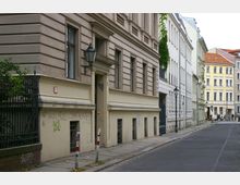 Straßenszene mit alten Wohngebäuden, darunter ein sandfarbenes Gebäude mit verzierten Fassaden und Graffiti an der unteren Wand. Auf dem Bürgersteig stehen Metallpoller, eine Laterne und ein schmiedeeisernes Tor; im Hintergrund weitere mehrstöckige Gebäude mit teils bunten Fassaden.