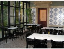 Ein Café- oder Restaurantinnenraum mit schwarzen Holzstühlen und weißen Tischen auf einem Boden aus Steinfliesen. Eine Wand zeigt ein geometrisches Muster aus blau-weißen Kacheln, während eine andere Seite von großen Glasfenstern mit Blick auf Bäume geprägt ist; im Hintergrund sieht man eine hölzerne Tür mit einem verzierten Steinrahmen.