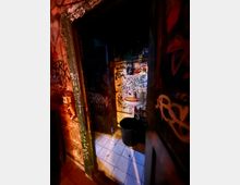 Schmaler, schwach beleuchteter Toilettenraum mit weißen Bodenfliesen und einem kleinen Waschbecken an einer gefliesten Wand. Wände und Türrahmen sind dicht mit Graffiti und Aufklebern bedeckt, daneben steht ein schwarzer Mülleimer.