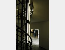 Ansicht eines modernen Gebäudeinnenraums mit einer Wendeltreppe aus Metallgeländern, einer beleuchteten Glaswand mit vertikalen Leuchtstoffröhren und einem Durchgang zu einem offenen Bereich mit glatten Marmorböden.