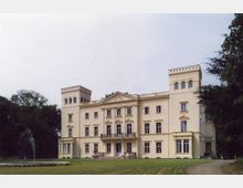 Helles Schloss im klassizistischen Stil mit zwei Seitentürmen und einem Mittelrisalit, umgeben von einer Rasenfläche und Bäumen; im Vordergrund ein Springbrunnen.