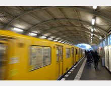 Eine U-Bahn-Station mit einem gelben U-Bahn-Zug, der am Bahnsteig hält. Der Bahnsteig hat eine gewölbte Metalldecke mit Beleuchtung, und Menschen gehen entlang des Gehwegs in Richtung des Zuges.