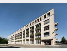 Historisches Industriebauwerk mit heller Backsteinfassade, großen Fensterreihen und grünen Fensterläden, aufgeteilt in mehrere Etagen; modernistischer Architekturstil. Das Gebäude steht auf einem gepflasterten Hof, umgeben von Bäumen und klarem Himmel.