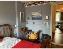 Ein gemütliches Schlafzimmer mit blauen Wänden, einem hölzernen Bettgestell, einem kleinen Schreibtisch aus Holz und einem Rattanstuhl. An der Wand hängen Bilder und unter einem Regal mit Büchern befinden sich Schreibmaterialien; im Hintergrund ist ein Blick in ein weiteres Zimmer erkennbar.