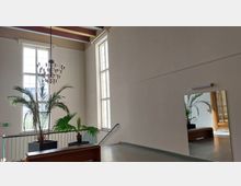 Heller Innenraum mit hohen Decken, mehreren großen Fenstern, einer hängenden Deckenleuchte und dekorativen Palmen in schwarzen Pflanzgefäßen. Ein Spiegel reflektiert den Raum, und im Hintergrund ist eine Holztür zu sehen.