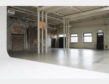 Large industrial-style indoor studio or gym space with a smooth concrete floor, exposed brick and painted walls, and tall support columns under an open ceiling with beams and ductwork. Two basketball hoops are mounted on the wall and a column, with black doors and rectangular windows along one side.