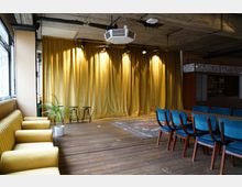 Ein kleiner Veranstaltungsraum mit Holzboden, einer Bühne mit goldenen Vorhängen und zwei schwarzen Hockern. Im Vordergrund befinden sich gelbe Polstersessel, ein grüner Pflanzenkübel und blau gepolsterte Stühle um einen Holztisch.