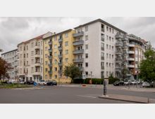 Straßenecke mit mehrstöckigen Wohnhäusern verschiedener Fassadengestaltungen: links elfenbeinfarbene Putzfassade, in der Mitte gelbe Kacheln mit Balkonen und rechts weiße Kachel- sowie eine braun-weiße Rundfassade. Vor den Gebäuden stehen parkende Autos, Straßenschilder und vereinzelte Bäume.