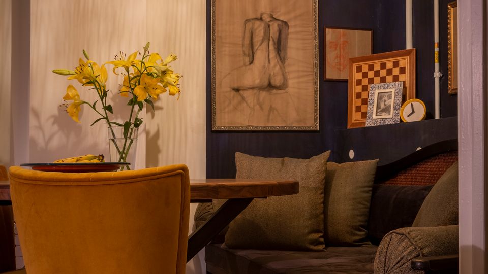 Gemütliche Sitzecke in einem Café oder Wohnraum mit dunkler Eckbank und Kissen, Holztisch und senfgelben Polsterstühlen auf gemustertem Fliesenboden. An der dunkelblauen Wand hängen gerahmte Bilder, darüber eine große runde Pendelleuchte; auf dem Tisch steht eine Vase mit gelben Blumen.