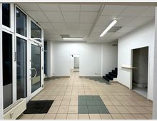 Ehemaliges Ladenlokal mit beige gefliesten Böden und weiß gestrichenen Wänden. Große Glasfront mit Doppeltür auf der linken Seite, eine Treppe mit dunklem Belag führt in einen erhöhten Bereich, und eine seitliche Tür führt in einen kleinen Nebenraum.