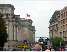 Straßenszene in Berlin mit dem Reichstagsgebäude im Hintergrund, erkennbar an seiner historischen Architektur und der deutschen Flagge auf dem Dach. Im Vordergrund eine belebte Straße mit Autos, Fußgängern und einem Touristenbus.